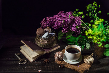 White cup with coffee on a linen napkin and old wooden table and stack of macarons. Old book and lilac.