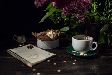 Coffee and cream macaroons in a white plate with coffee cup, eco notebook with a pencil under lilac branch.