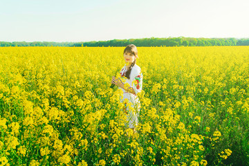 Fototapeta premium Beautiful young woman in Ukrainian embroidered standing in a field of yellow rape flowers