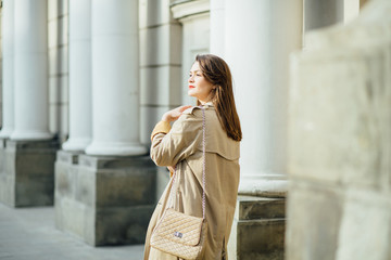 Slim elegant brunette woman with red lips in a beige coat and black skirtwith long legs near the columns.