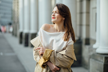 Slim elegant woman with red lips in a beige coat and black skirt brunette with long legs near the columns.