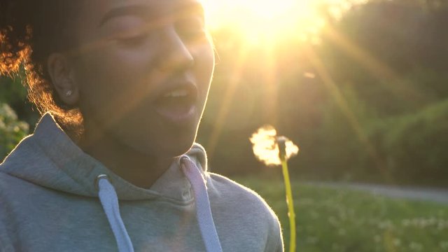 4K video clip of beautiful healthy mixed race African American girl teenager girl young woman laughing, smiling and blowing a dandelion at sunset or sunrise