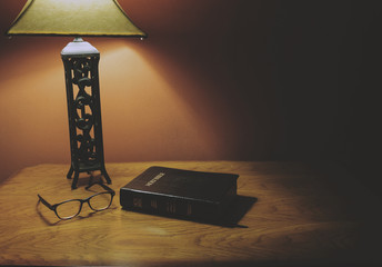 Holy Bible Lying On A Wood Table With Glasses And Lamp Shining.
