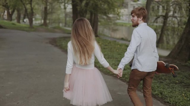 Young couple in love walking in summer park holding hands. Woman in pink tutu skirt man with longboard in hands Three-axis Gimbal stabilized tracking shot