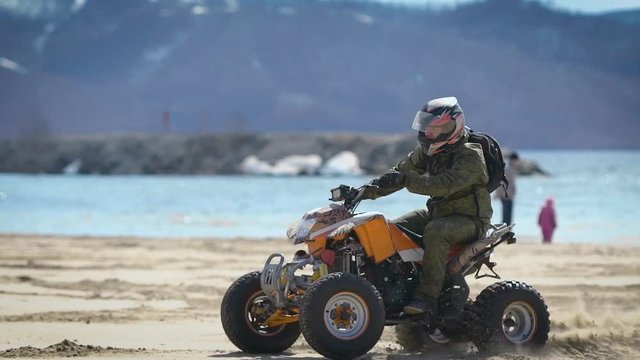 Close up of man in protective equipment and helmet driving his ATV and moving along the shore with mountains in the background. Extreme sportsman riding his quad bike on the beach with nature behind.