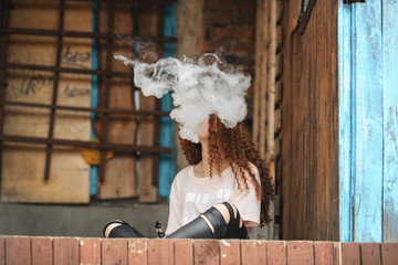 Beautiful vape teenager. Portrait of a pretty young white girl with red curly hair vaping an electronic cigarette near the old ruined village house. Lifestyle.