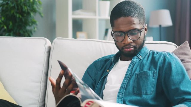 Young african man in glasses, blue shirt sitting on white sofa viewing magazine for shopping. Indoor.
