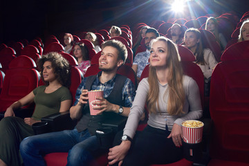 Smiling positive couple of boyfriend and blonde girlfriend watching funny movie at cinema . Best friends spending time together, eating popcorn when looking at project. Big fiends group.