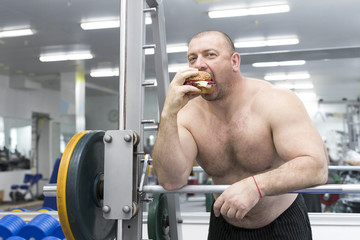 Big fat hungry man chewing a hamburger with meat and cheese in the gym 