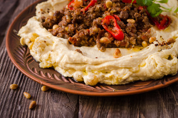 Creamy hummus with spicy mutton minced meat in clay bowl on wooden background. Ramadan food.
