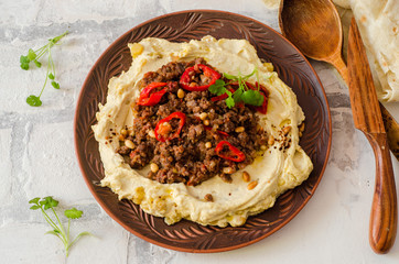 Creamy hummus with spicy mutton minced meat in clay bowl on stone background. Ramadan food. Top view