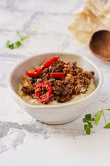 Creamy hummus with spicy mutton minced meat in clay bowl on stone background. Ramadan food.
