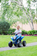 Portrait of smiling little Caucasian boy toddler driving blue electric car on the road path outside on spring summer day, seasonal child activity concept, healthy childhood lifestyle