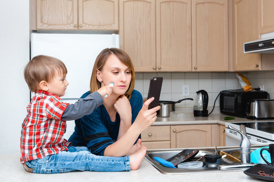 White Caucasian young woman mother housewife taking pictures selfie in kitchen, her child son boy playing and drawing on her cheek with marker, crazy parent life, funny kid moment