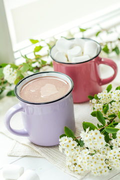 Hot cocoa with marshmallows in pink cups and fresh spring white flowers on the windowsill. Cozy home concept. Shallow depth of field.