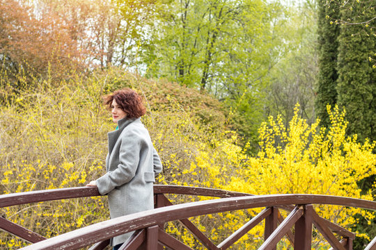 Pretty young red head woman on the bridge in spring park. Green trees and forsythia blossoms background.