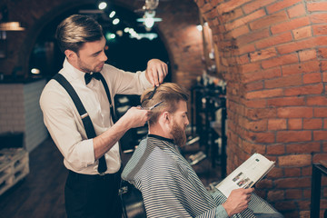 Red bearded guy is reading a magazine in a barber shop while getting bren new haircut from a classy dressed stylist. Both are buzy