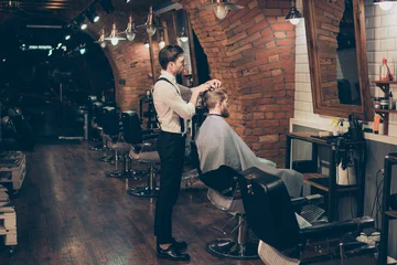 Foto auf Acrylglas Friseur Barber shop classy dressed specialist is styling the hair of a client. Salon is retro and vintage. Customer is a young bearded man, covered with cape