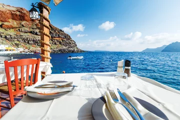 Rolgordijnen Served table with white tablecloth against blue water of Aegean sea on Santorini island resort in Greece, Europe. © Feel good studio