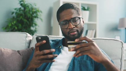 Young african man sitting on the couch, using smart phone for online shopping with credit card on living backround. Indoor. - Powered by Adobe