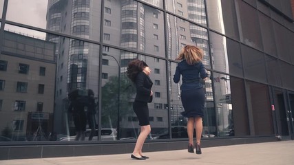 two girls in the clothing business is happy, jumping and having fun. business woman celebrating success and victory. businessmen on background of office building of glass. slow motion - Powered by Adobe