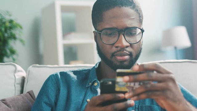 Potrait of attractive young african man with glasses using smart phone for shopping with credit card at living room. Indoor.