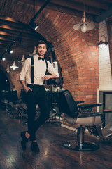 Handsome classy dressed stylist in a barber shop. He is successful and smiling, standing with crossed legs in a retro designed barbershop