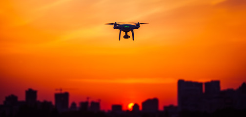 Fototapeta na wymiar Modern Remote Control Air Drone Fly high with action camera in sunset sky. Cityscape silhouette in the background. Modern technologies. Travel, hobby, inspiration. Orange toning filter. Kiev, Ukraine