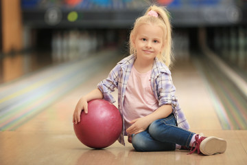 Cute little girl with ball sitting on floor in bowling club