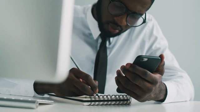 Portrait of young african man working in office, making some calculations on the smart phone and writing result in note book.