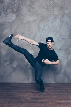 Handsome young break dancer is making his favourite move in studio on the grey background. He is wearing black stylish outfit and cap