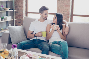 True love. Beautiful couple in casual outfit is sitting on modern sofa and smiling. They are drinking mugs of tea and look at each other with tenderness