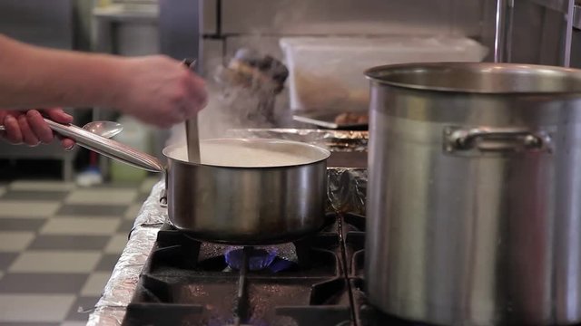The chef cooks white rice in stewpan on the kitchen of restaurant. The cook mixes up rice in stainless steel saucepan with metal slice. Dish prepares on the black gas stove. Grain rice boil in enough
