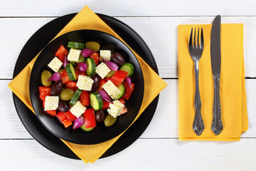 delicious Greek salad with fresh vegetables, feta cheese green and kalamata olives, red onion bulbs on black plates on white boards with fork and knife on napkin, view from above