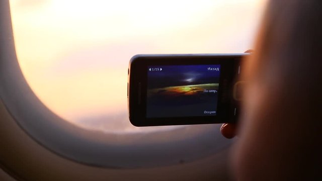 Closeup of mobile phone in hands of cute funny kid taking pictures of beautiful sunset sky in plane during flight. Child of 9 years sits on his seat near window. Real time full hd video footage.