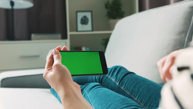Woman hands using smart phone with green screen for chating on skype while relaxing on the sofa in the living room. Chroma key. View from the back. Horizontal position.