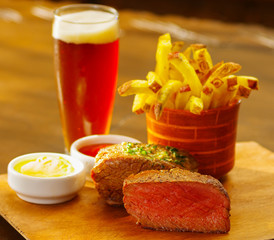 Well-done grilled marinated beef flank steak with ketchup, mustard, french fries and a glass of beer on wooden board