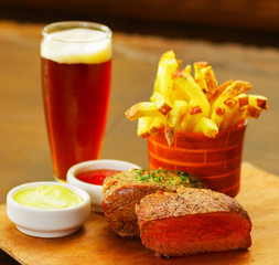 Close up of a well-done grilled marinated beef flank steak with ketchup, mustard and french fries with a glass of beer on wooden board