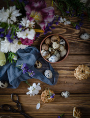 quail eggs and walnuts in a brown plate with cookies, white and purple flowers, blue cloth, scissors on a brown wooden table