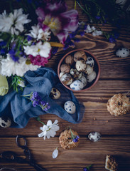 quail eggs and walnuts in a brown plate with cookies, white and purple flowers, blue cloth, scissors on a brown wooden table toned for a vintage effect