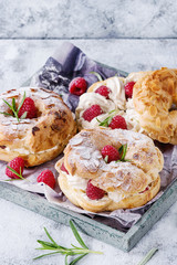 Homemade choux pastry cake Paris Brest with raspberries, almond, sugar powder and rosemary, served on wooden serving tray over gray blue texture background. French dessert. Close up