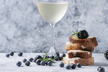 Grilled sandwich with melted goat cheese, blackberry, blueberry, rosemary and honey, served on wooden serving board with glass of cold white wine over gray blue texture background. Summer appetizer.