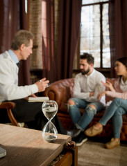 Young man and woman sitting on sofa and communicating with psychology at psychologist's office. Closeup of hourglass representing on table.