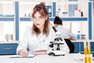 young scientist in lab coat working with microscope and writing in folder in chemical lab