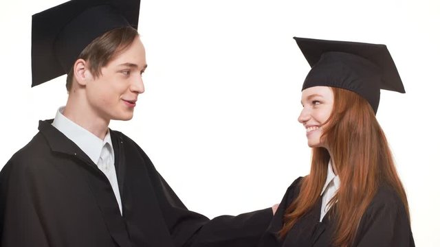 Happy young Caucasian graduate male and female in black robes and square academical caps laughing and talking with each other standing on white background
