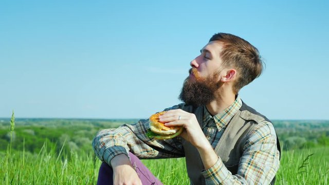 A young bearded man eating a hamburger outdoors. Sits on a meadow in a picturesque place In sring sunny day