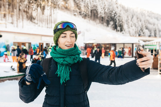 The attractive girl in winter clothes takes a selfie. She has flicked out tongue language and winks. Around her there are a lot of people.