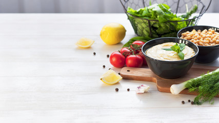 Bowl with hummus, chickpeas, lemon, cherry tomatoes and herbs on a white rustic wooden background. Middle Eastern cuisine. Background hummus with place for text.