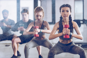 group of athletic young people in sportswear with dumbbells squatting and exercising at the gym