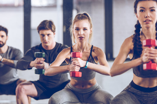 group of athletic young people in sportswear with dumbbells squatting and exercising at the gym
